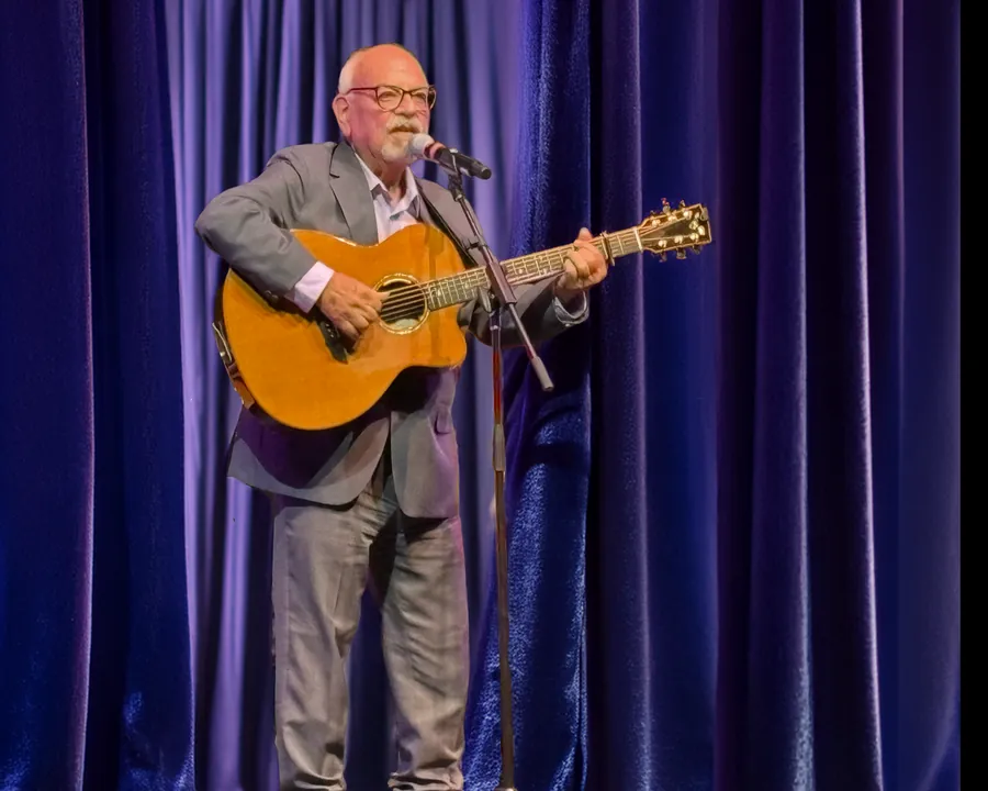 Ron singing with an acoustic guitar in front of a purple stage curtain